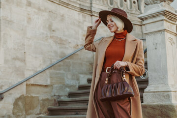 donna elegante nel colori di terra: trench beige, pantaloni, borsa e cappello marroni e dolcevita rosso mattone