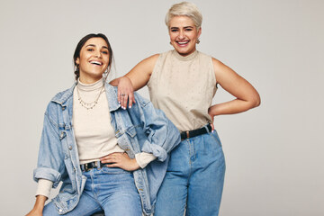 due donne sorridenti vestite in denim: una indossa jeans, un dolcevita beige e una giacca di jeans, l'altra solo jeans e camicetta beige